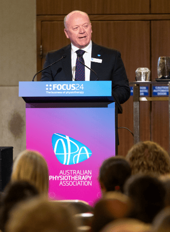 The head of the APA, Scott Willis standing at a podium speaking. The podium has a sign sayingFOCUS24.